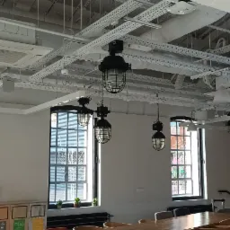 Wnętrze biura z industrialnym oświetleniem sufitowym, widoczne lampy w metalowych koszach, systemy wentylacyjne i białe rury pod sufitem, duże okna z czarnymi ramami.