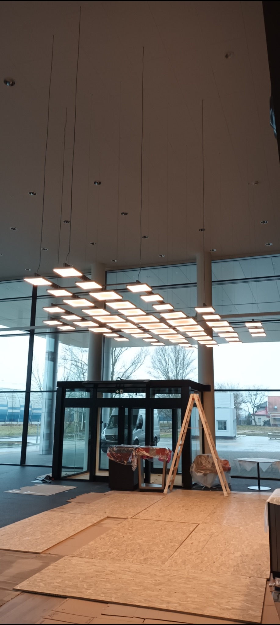 Sufit z kwadratowymi panelami LED w nowoczesnym biurze, widoczne przygotowania do remontu, zabezpieczona podłoga płytami OSB, drabina oparta o framugę drzwi, duże okna z widokiem na parking.