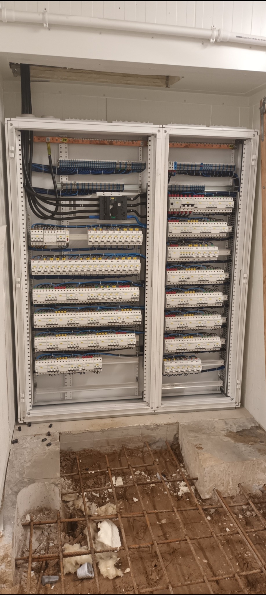 Otwarta szafa elektryczna z licznymi bezpiecznikami i przewodami, widoczna instalacja pod podłogą w trakcie prac wykończeniowych.