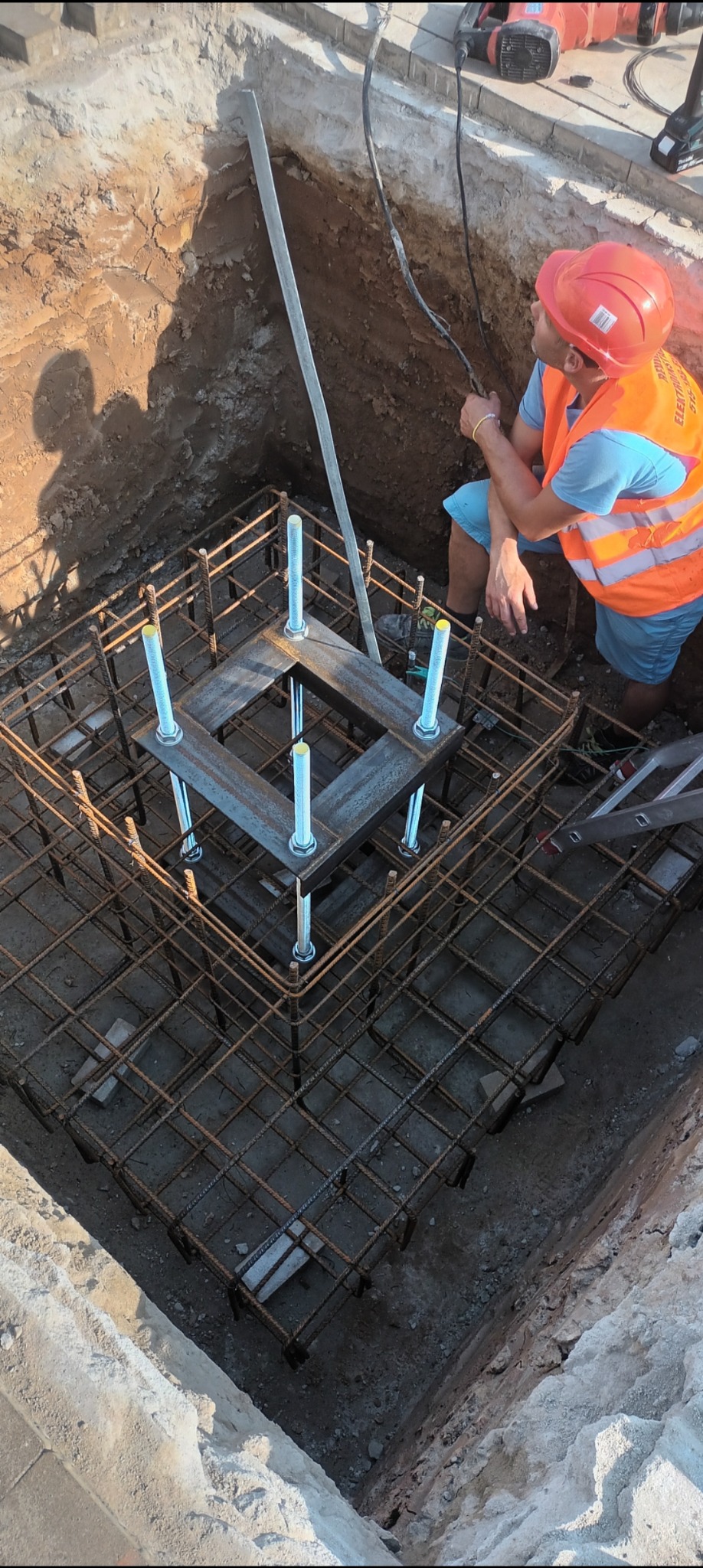 Pracownik w pomarańczowej kamizelce i kasku nad wykopem z konstrukcją zbrojeniową i metalowymi elementami mocującymi, widoczne narzędzia elektryczne w tle.