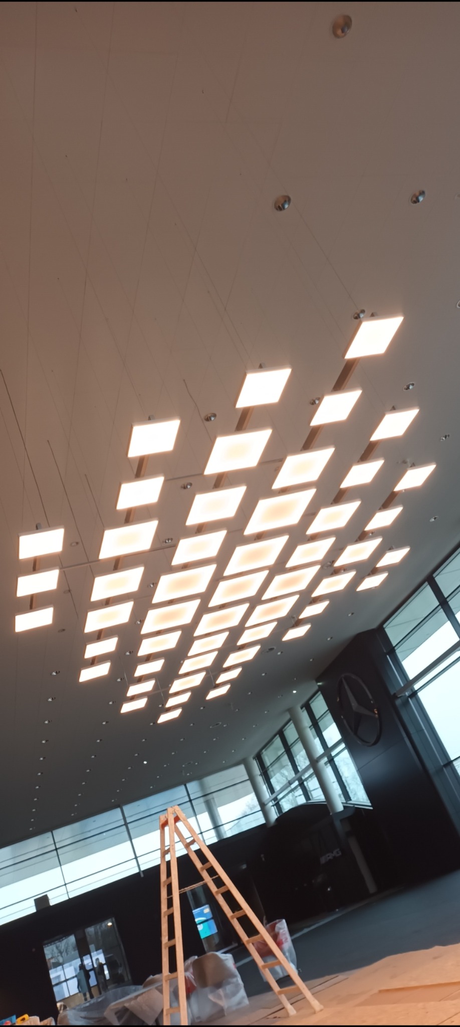 Sufit z rzędem kwadratowych paneli LED w salonie samochodowym, widoczna drabina i logo marki, ujęcie z dołu.