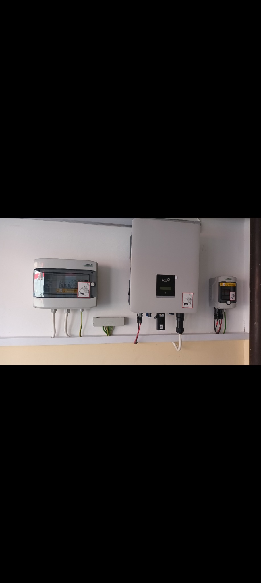 Rozdzielnica elektryczna, inwerter fotowoltaiczny i licznik energii zamontowane na ścianie, z widocznymi przewodami i oznaczeniami PV.