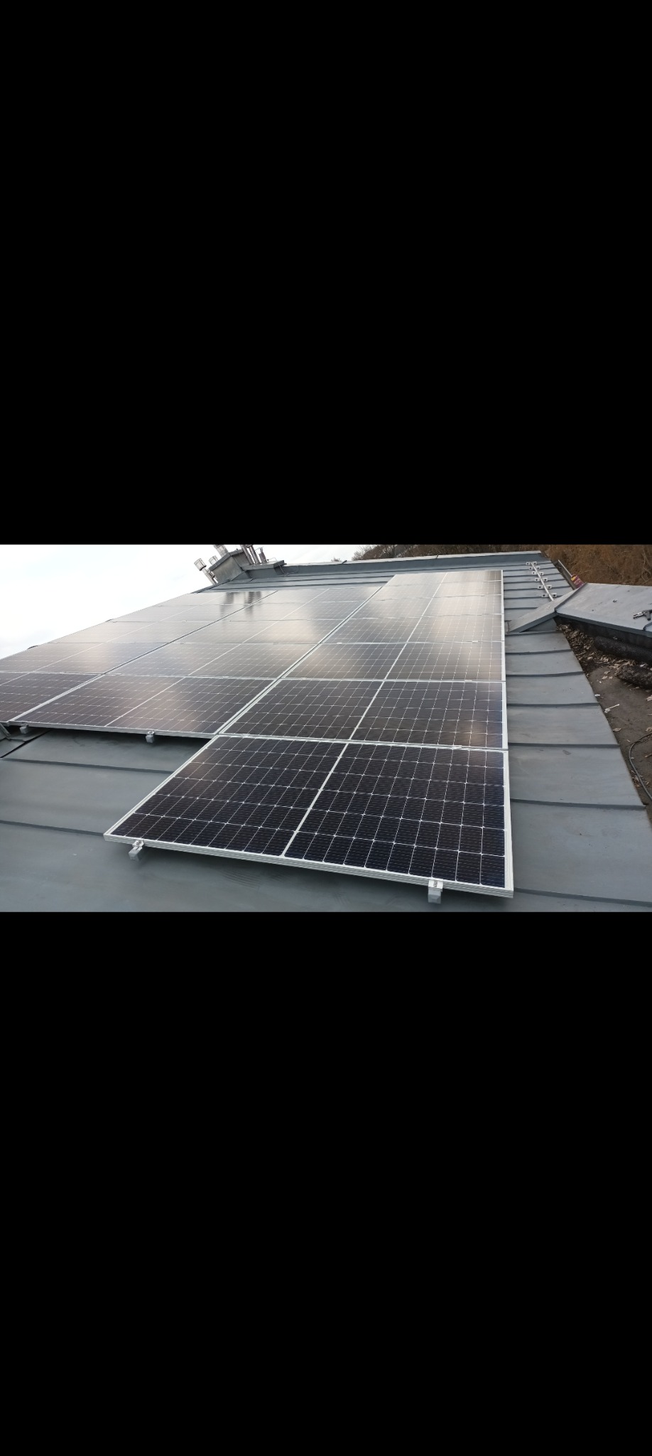 Panele fotowoltaiczne zamontowane na szarym, metalowym dachu, widoczne z perspektywy ukośnej, z elementami systemu mocowania i drabinką kominiarską w tle.