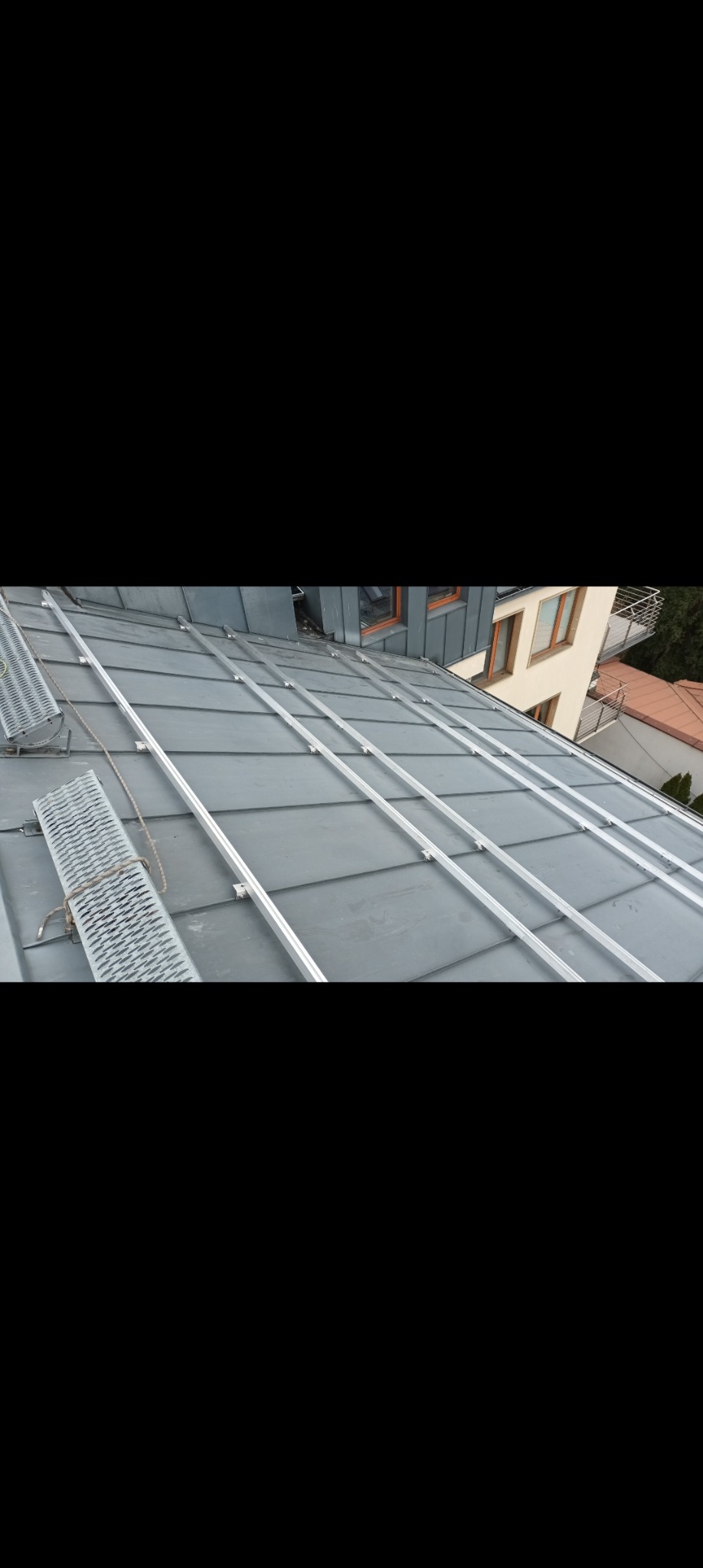 Szary dach z blachy trapezowej z zamontowanymi aluminiowymi szynami, widoczne elementy mocujące, w tle budynki mieszkalne.