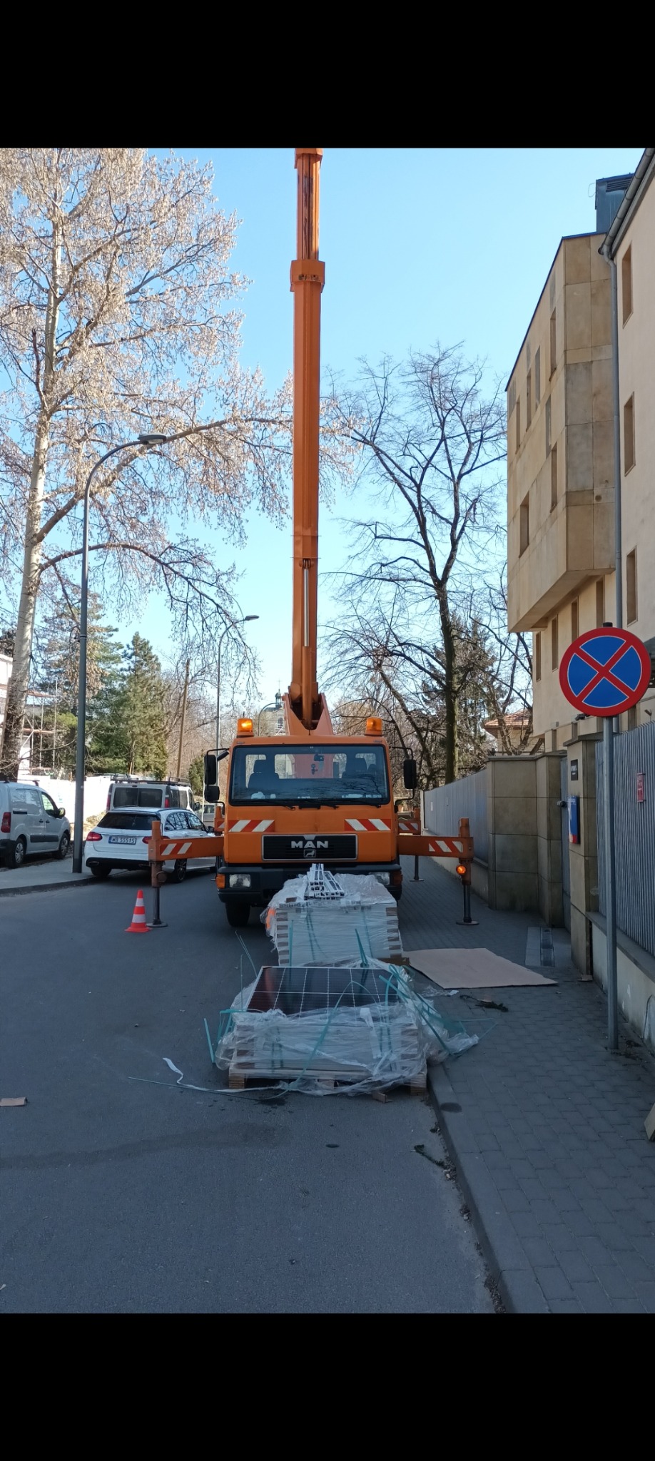 Pomarańczowy dźwig MAN z podnośnikiem teleskopowym na ulicy, przygotowany do montażu paneli fotowoltaicznych, widoczne palety z panelami w folii, znak zakazu parkowania.