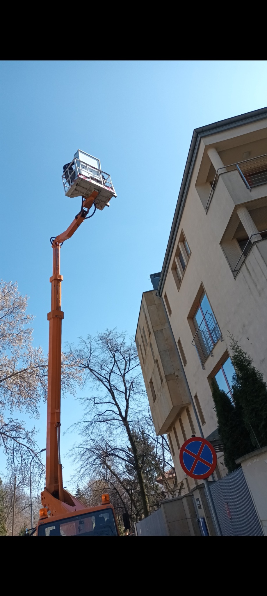 Pomarańczowy podnośnik koszowy z pracownikiem na wysokości przy budynku mieszkalnym, błękitne niebo w tle.