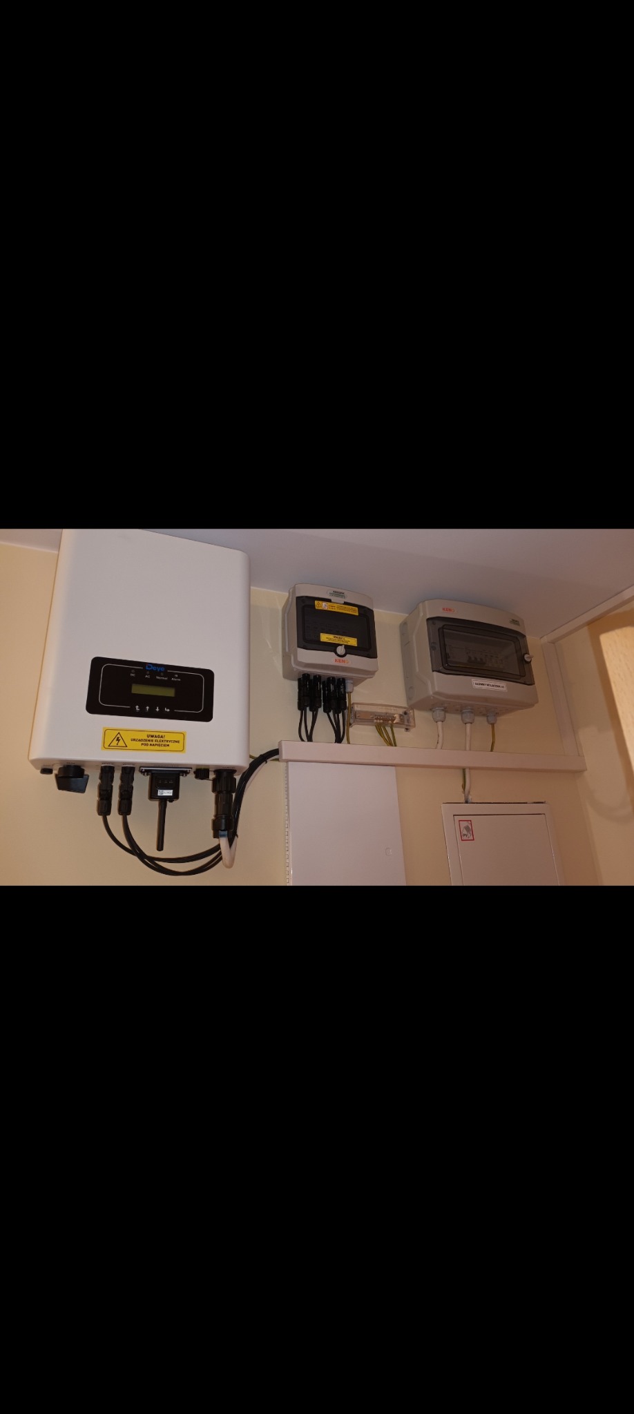 Biały inwerter fotowoltaiczny Devo z podłączonymi kablami, obok dwie szare skrzynki elektryczne na ścianie wewnątrz budynku.