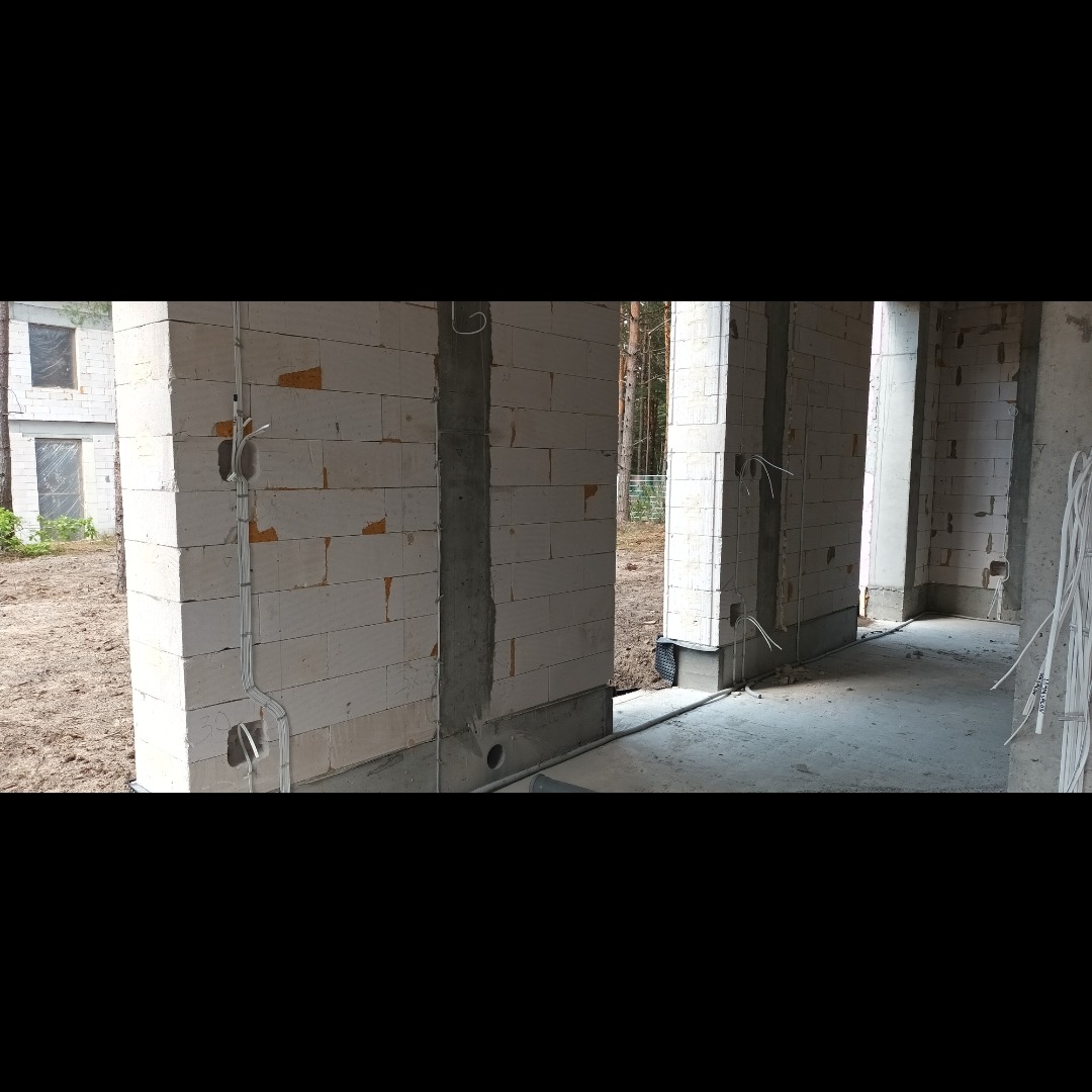 Wewnętrzne ściany z betonu komórkowego w trakcie budowy, z widocznymi przewodami elektrycznymi w peszlach, przygotowanymi do instalacji gniazdek i włączników, na tle lasu widocznego przez otwory...