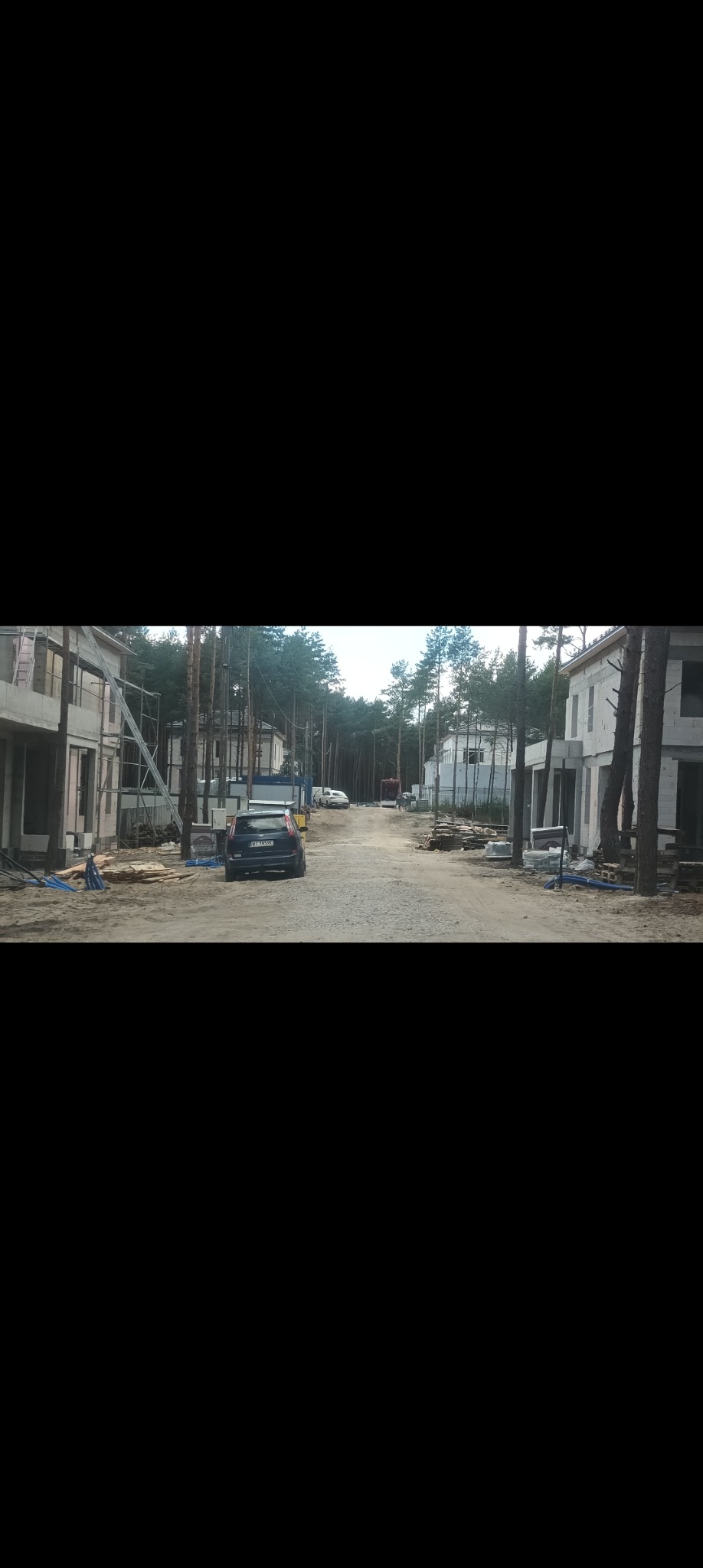 Nowe budynki w stanie surowym zamkniętym, z widocznymi instalacjami elektrycznymi na zewnątrz, otoczone lasem sosnowym i piaskową drogą dojazdową, z zaparkowanym samochodem osobowym.