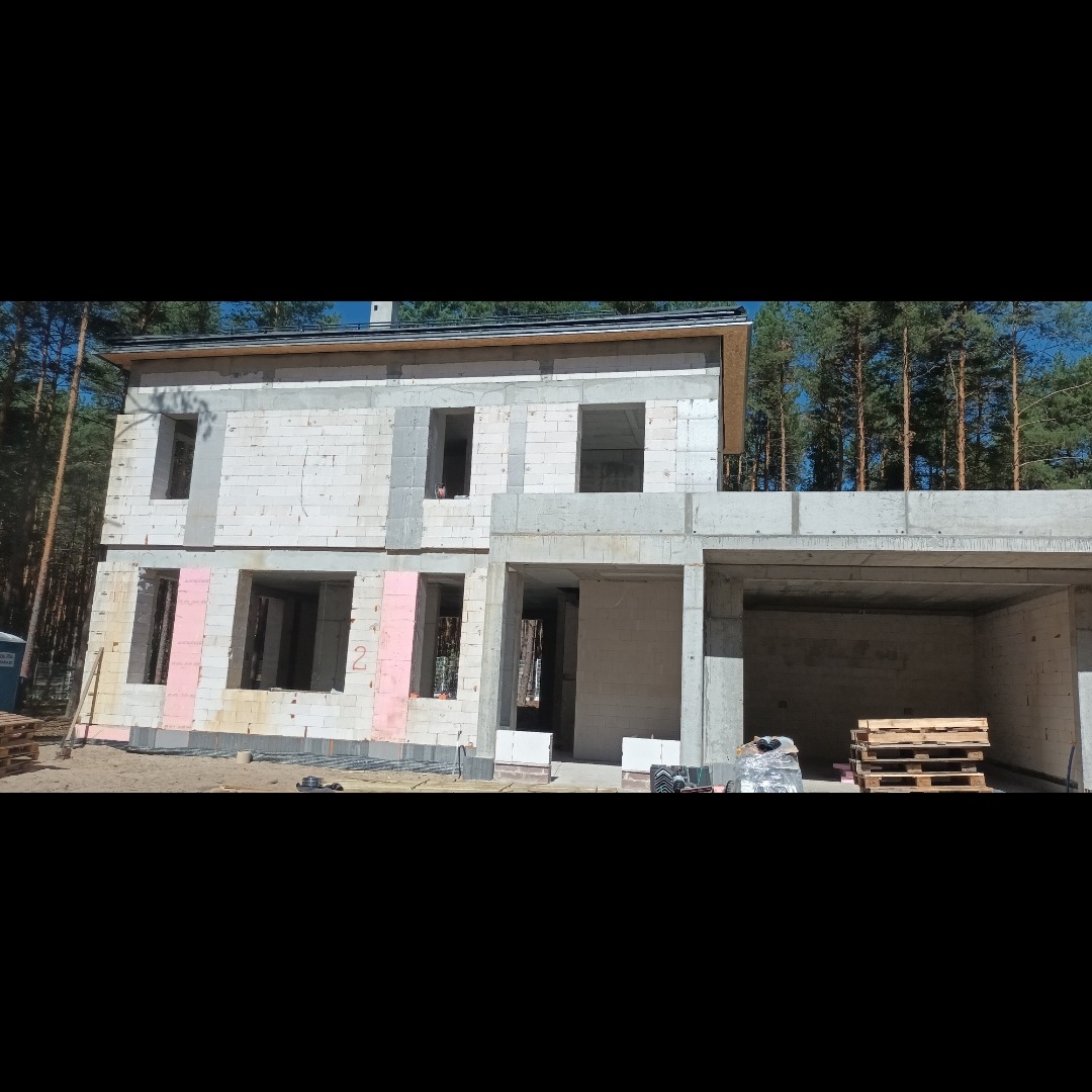 Surowy stan dwukondygnacyjnego domu z garażem w trakcie budowy, widoczne bloczki i ocieplenie, las w tle.