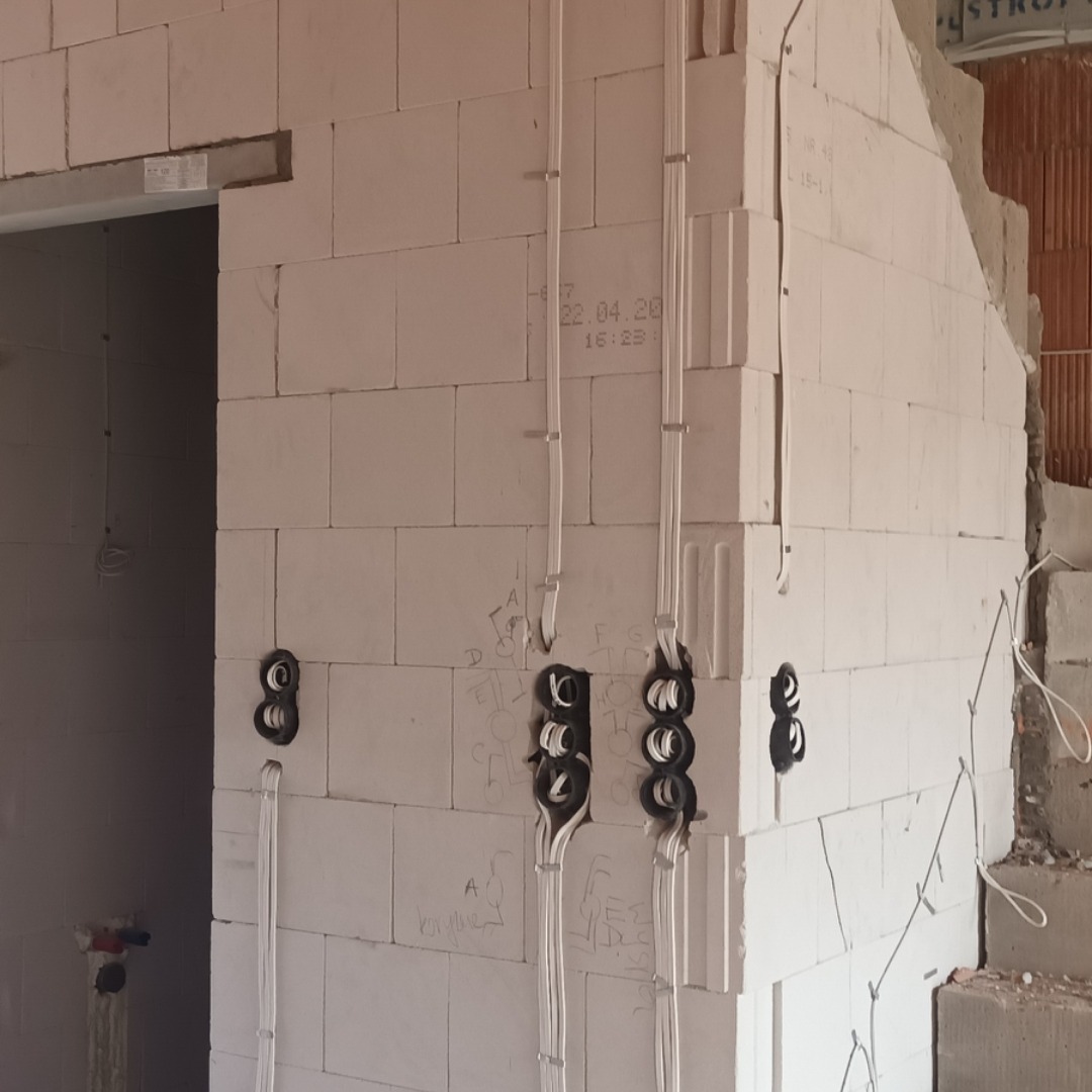 Surowy mur z betonu komórkowego z widocznymi puszkami elektrycznymi i przewodami w trakcie montażu, oznaczenia ołówkiem na ścianie.