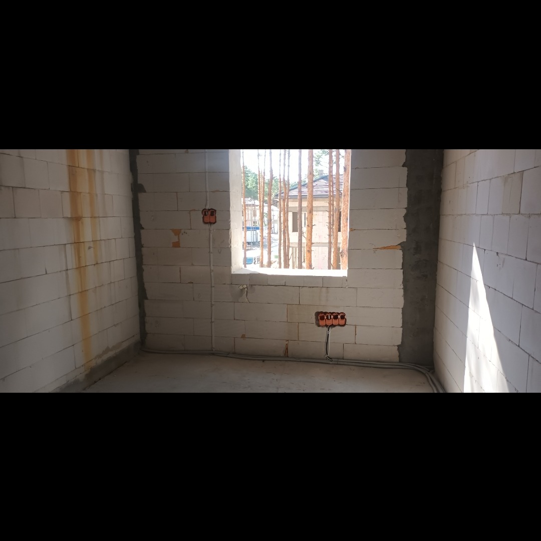 Surowy stan wnętrza z białych bloczków z widocznymi puszkami elektrycznymi i przewodami, okno z widokiem na drzewa.