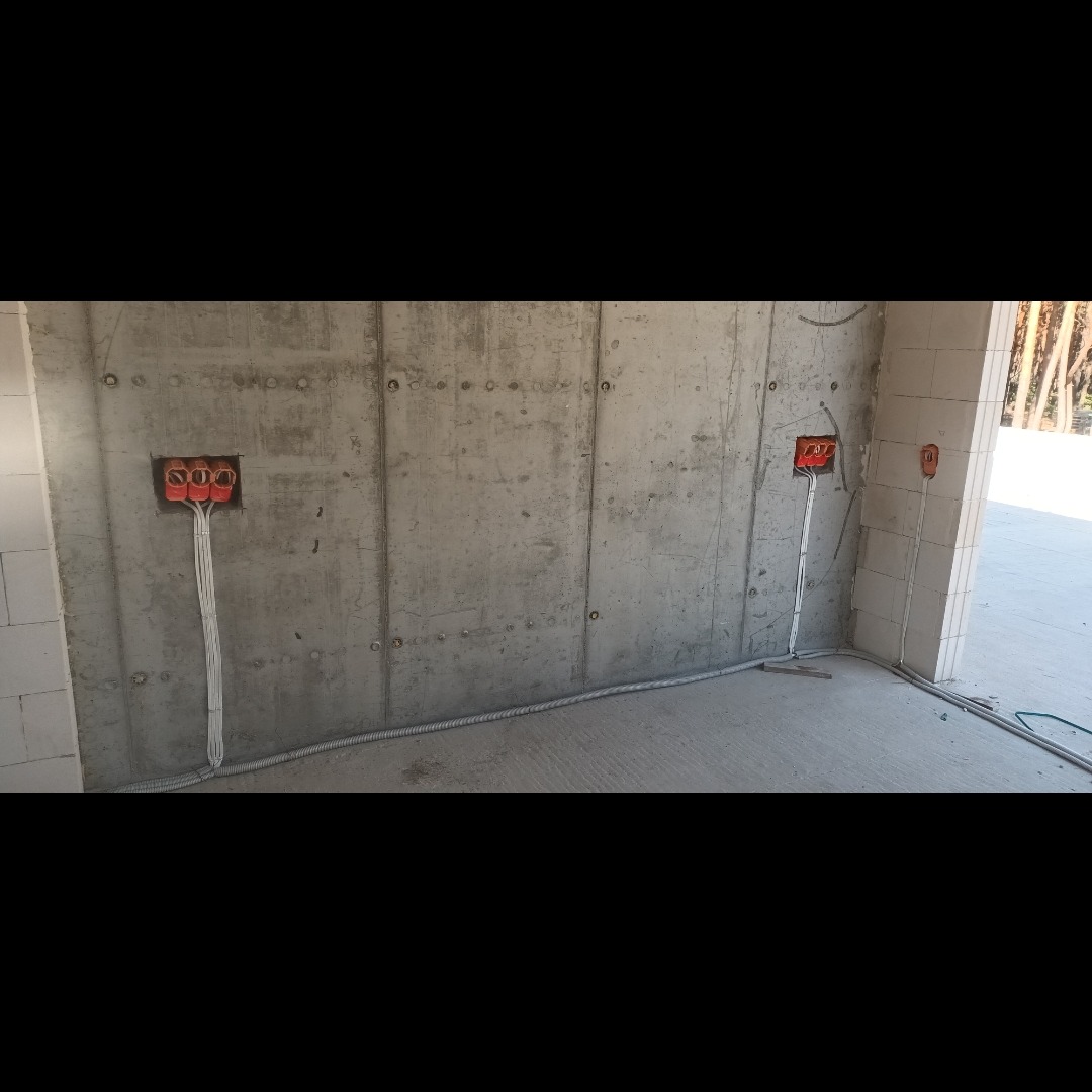 Surowa ściana z betonu z widocznymi puszkami elektrycznymi i przewodami w peszlu, instalacja w trakcie montażu.