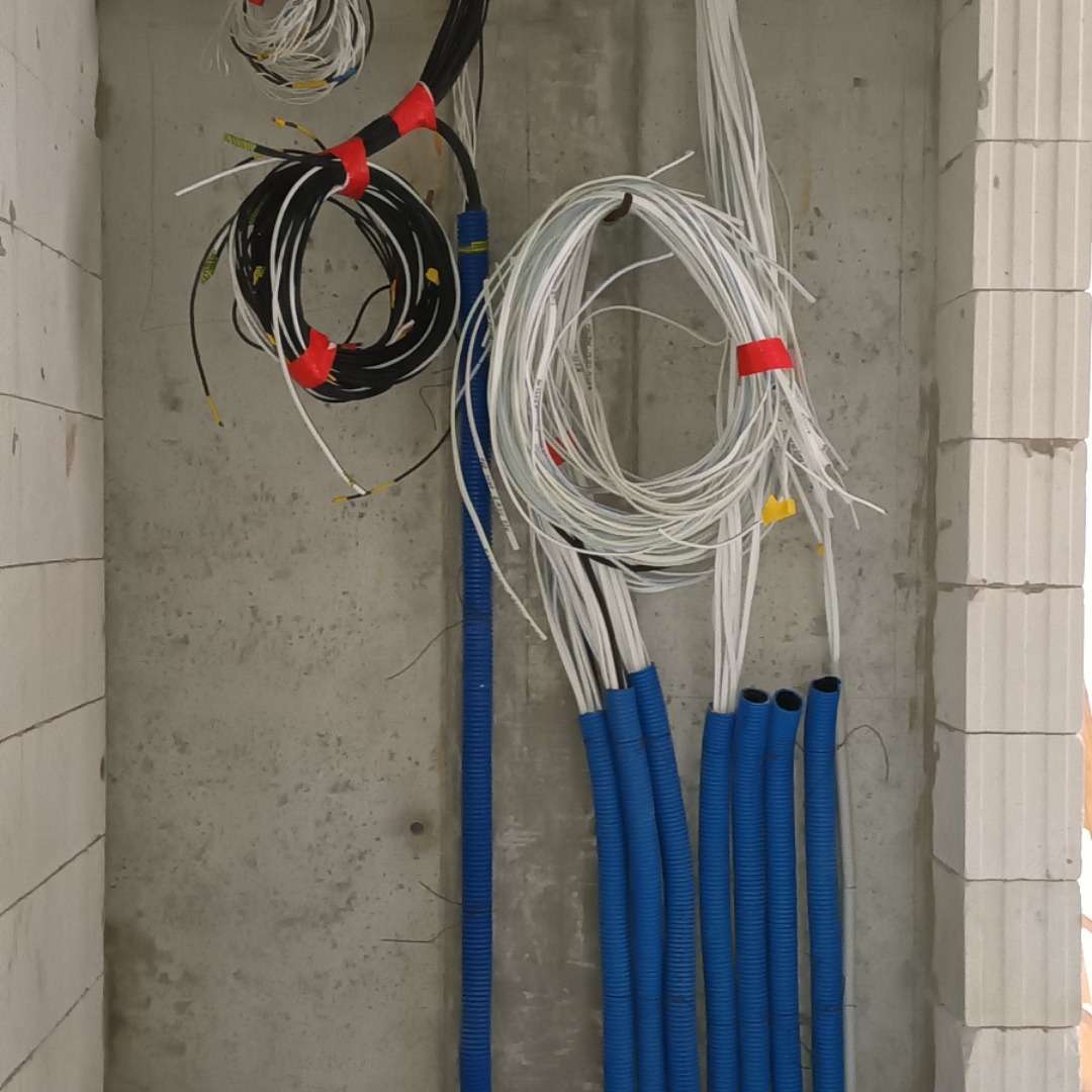 Wiązki kabli elektrycznych w niebieskich rurach karbowanych, przymocowane do surowej ściany z betonu komórkowego, zabezpieczone czerwoną taśmą izolacyjną.