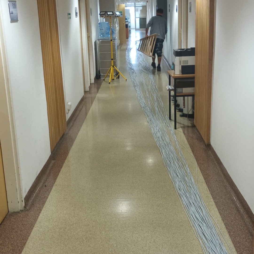 Długi korytarz biurowy z rozwiniętą wiązką szarych kabli elektrycznych na podłodze, pracownik w oddali niesie drabinę, widoczny sprzęt oświetleniowy i biurowy.