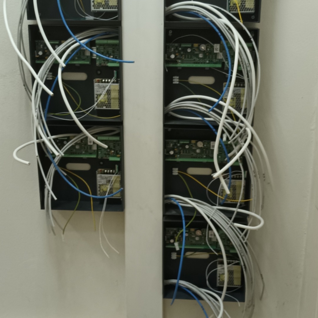 Dwa rzędy czarnych skrzynek z elektroniką i licznymi kablami (białe, niebieskie, żółte) wychodzącymi z nich, zamontowane na białej ścianie.