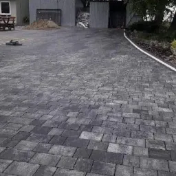 Ułożona, szara kostka brukowa na podjeździe z białą obwódką, z zagęszczarką w tle i przygotowanym miejscem na trawnik.