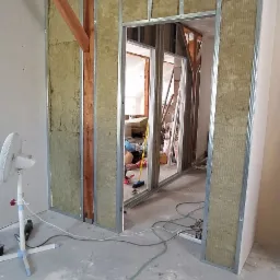 Montaż wełny mineralnej w metalowym stelażu pod płytę karton gips, widoczny fragment drewnianej belki stropowej i narzędzia na podłodze.