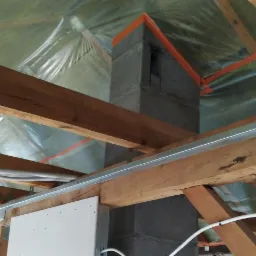 Konstrukcja drewniana poddasza z zamontowanymi profilami metalowymi, folią izolacyjną i fragmentem komina w trakcie prac wykończeniowych.