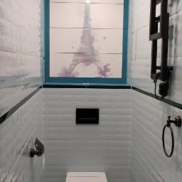 Wąska łazienka w jasnych odcieniach z toaletą wiszącą, grzejnikiem drabinkowym w kolorze ciemnego brązu i płytkami z motywem wieży Eiffla nad zabudową toalety.