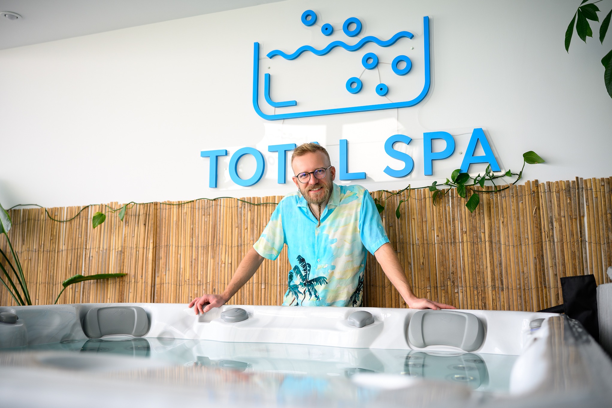 Uśmiechnięty mężczyzna w hawajskiej koszuli opiera się o brzeg wanny spa z hydromasażem. W tle logo firmy Total Spa i bambusowa ściana z zielonymi pnączami.