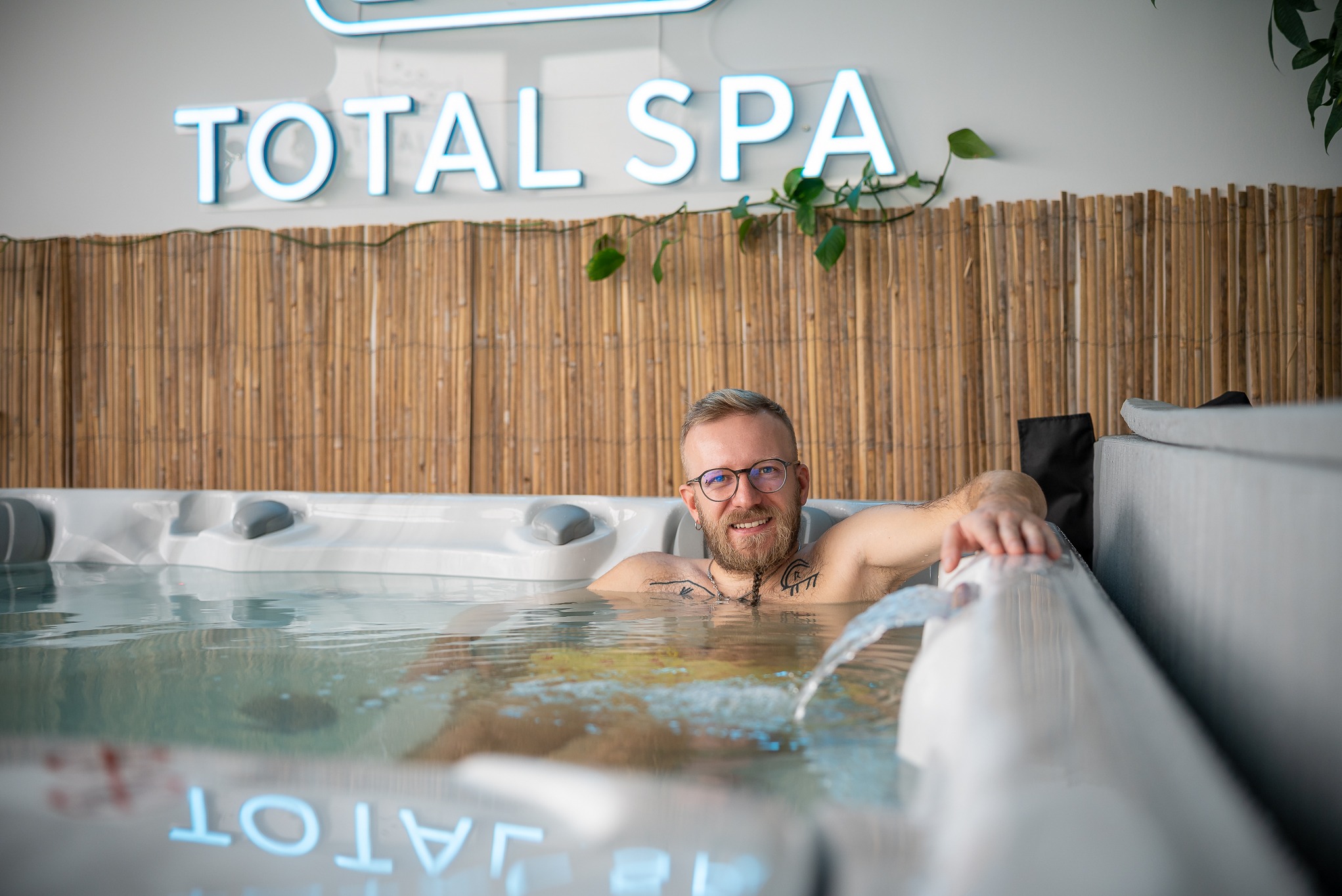 Mężczyzna w okularach relaksuje się w jacuzzi z hydromasażem. W tle bambusowa ściana i neon Total Spa. Woda w ruchu, widoczne dysze.