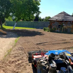 Glebogryzarka traktorowa przygotowuje ziemię pod trawnik na działce z altaną i betonowym ogrodzeniem w tle.
