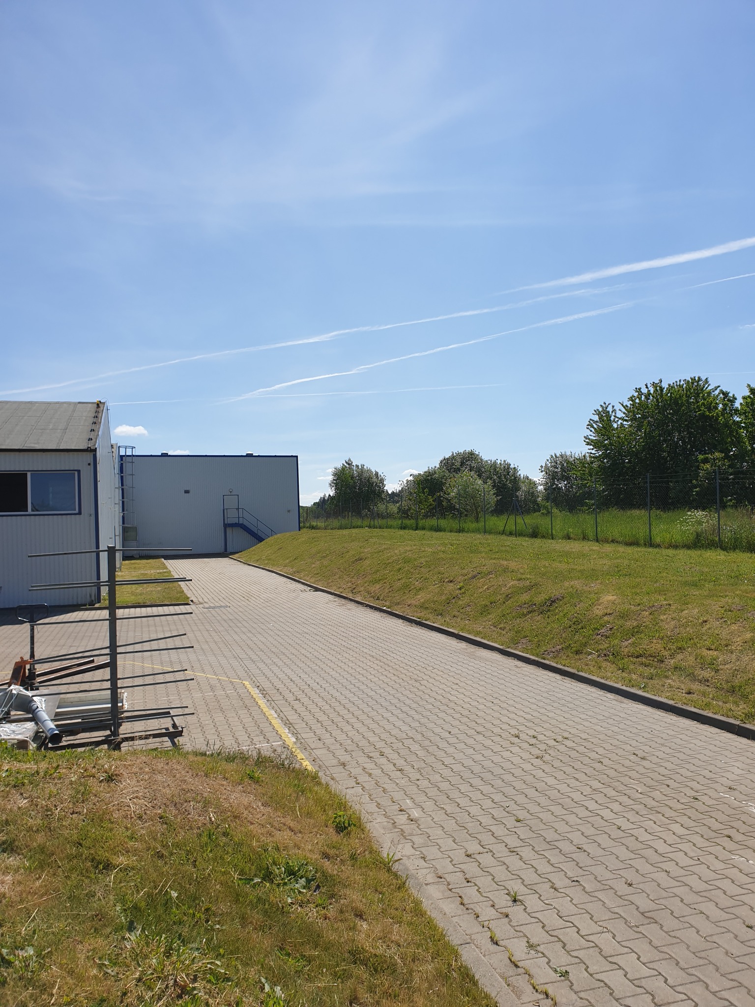 Widok z poziomu gruntu na budynek biurowy z równo przystrzyżonym trawnikiem, kostką brukową i błękitnym niebem z smugami kondensacyjnymi samolotów w tle. Lekkie zaniedbania w postaci uschniętej...