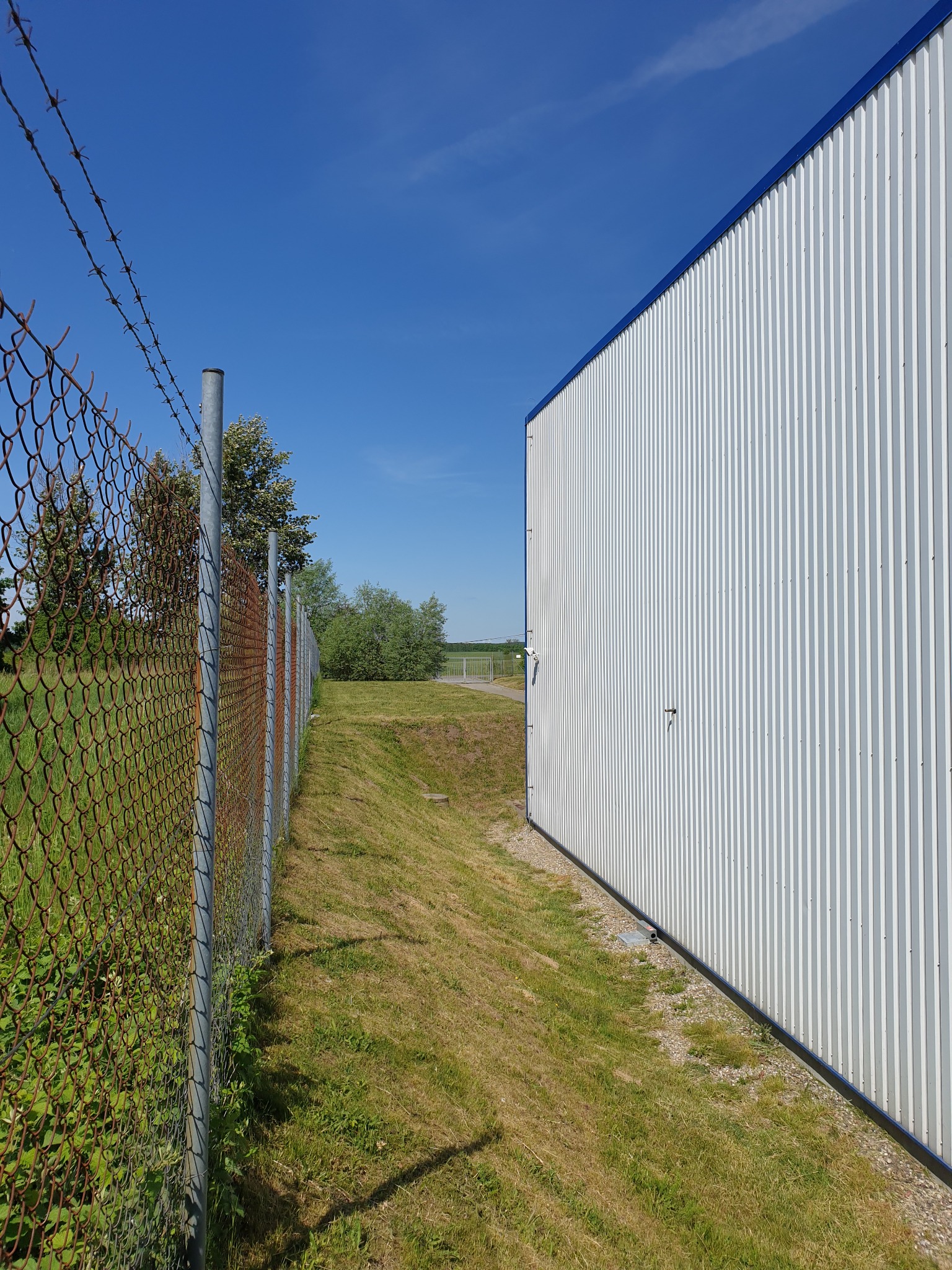 Teren przemysłowy z budynkiem o białej, blaszanej elewacji i ogrodzeniem z siatki z drutem kolczastym; trawa częściowo skoszona.
