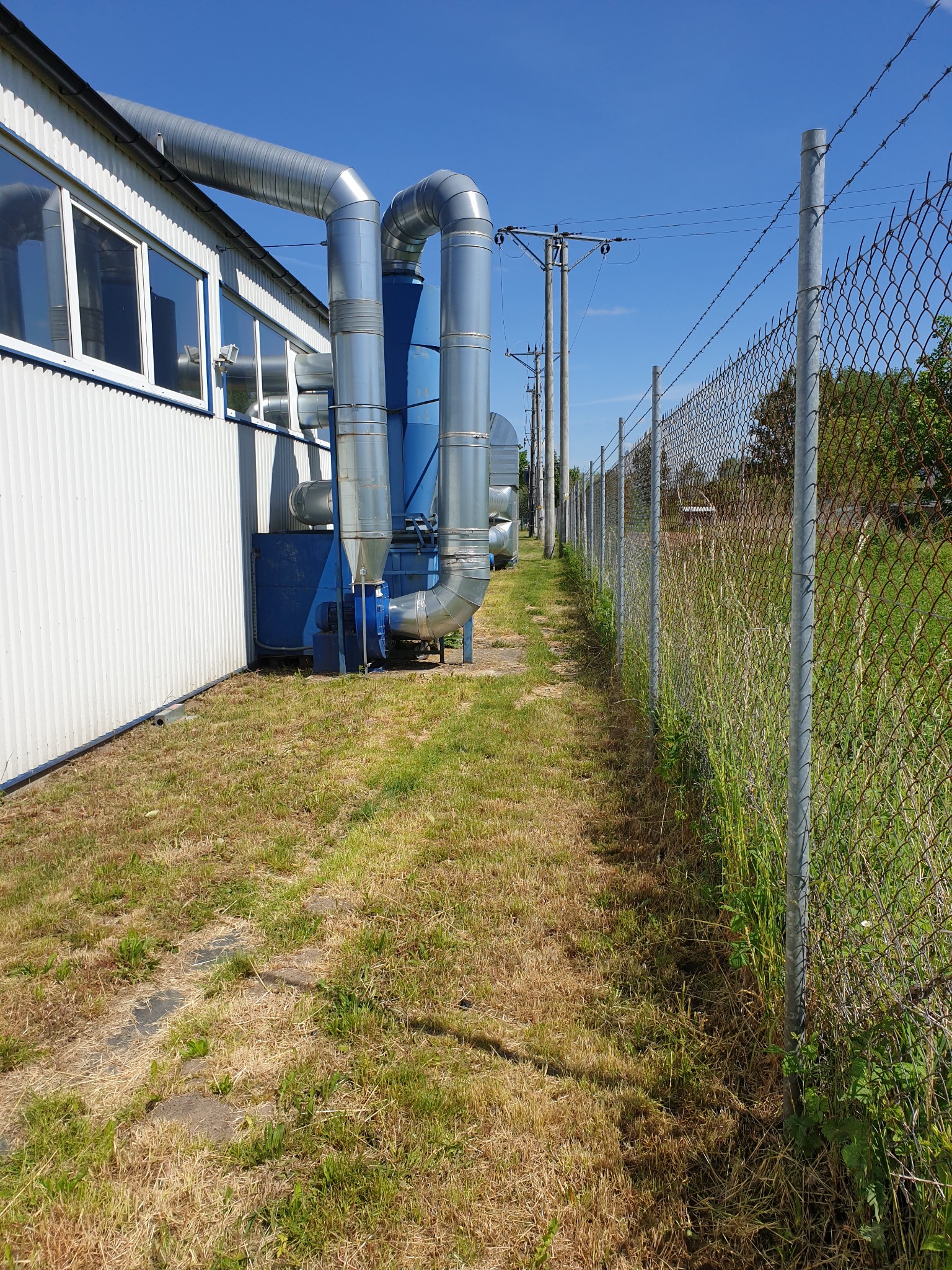 Widok instalacji przemysłowej z rurami wentylacyjnymi biegnącymi wzdłuż budynku i zaniedbanego trawnika przy ogrodzeniu z siatki.