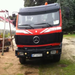 Czerwono-czarna ciężarówka Mercedes z widocznym logo na tle podwórza z zielenią i konstrukcją metalową.