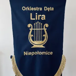 Granatowy sztandar z haftowanym złotym napisem 'Orkiestra Dęta Lira Niepołomice' i motywem liry, wykończony frędzlami. Precyzyjne wykonanie, elegancki wygląd.