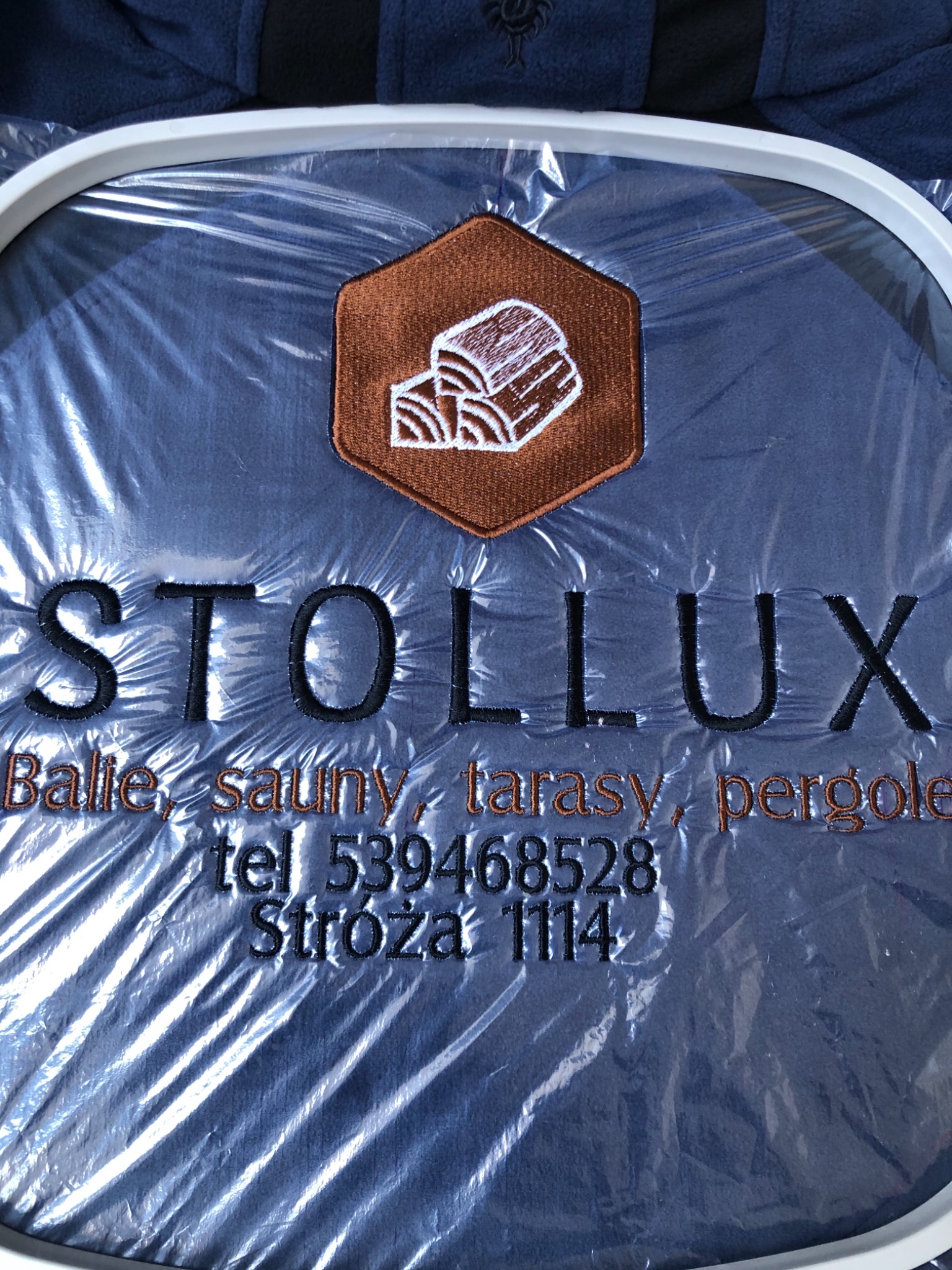 Haftowany logotyp firmy Stollux na granatowej tkaninie, z motywem drewna w brązowym sześciokącie, widoczne detale haftu i adres firmy.