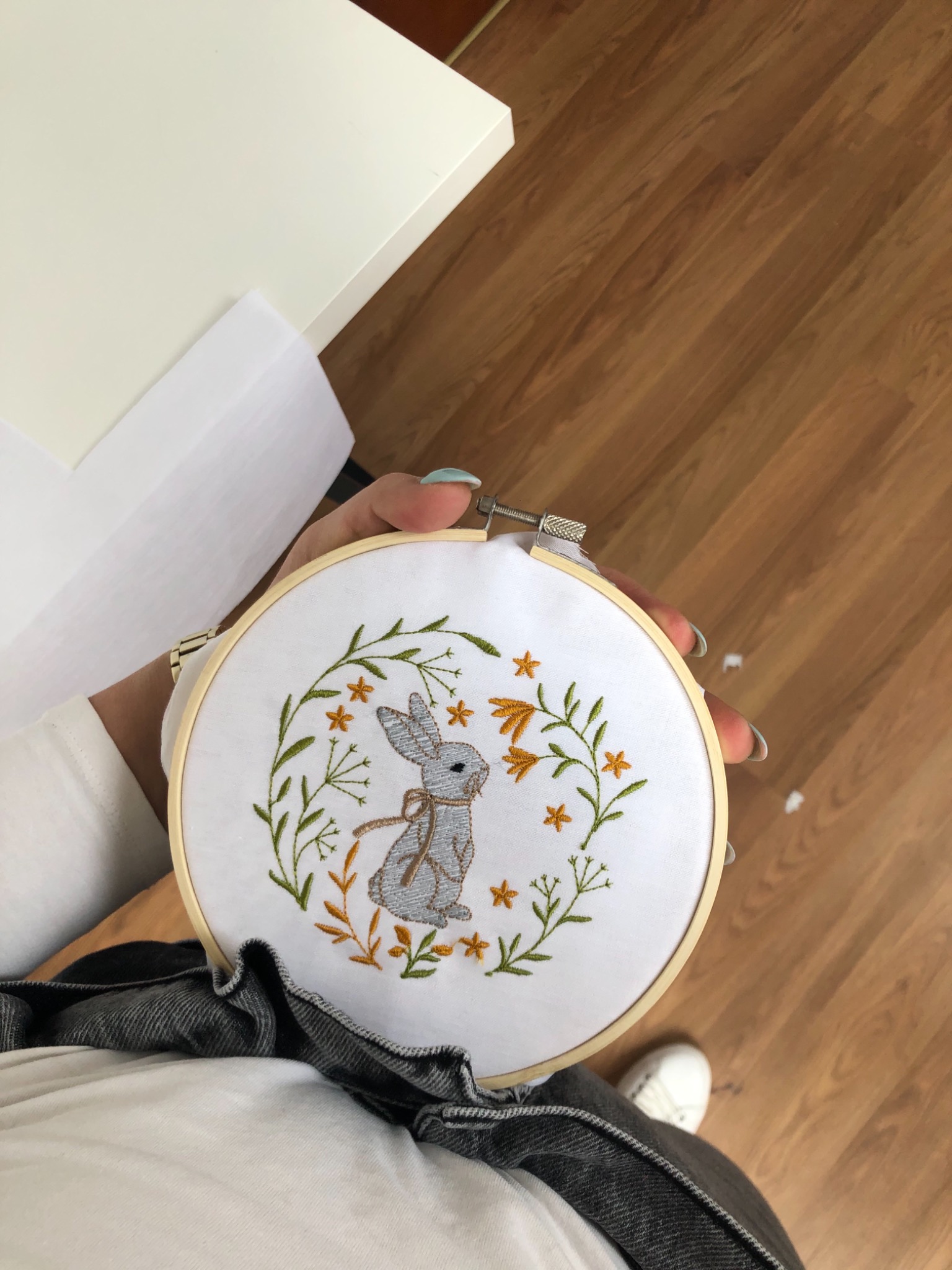 Ręcznie wykonany haft przedstawiający szarego zająca z kokardką, otoczonego roślinnym motywem w odcieniach zieleni i pomarańczy, umieszczony w drewnianym tamborku, trzymany w dłoni z błękitnym...