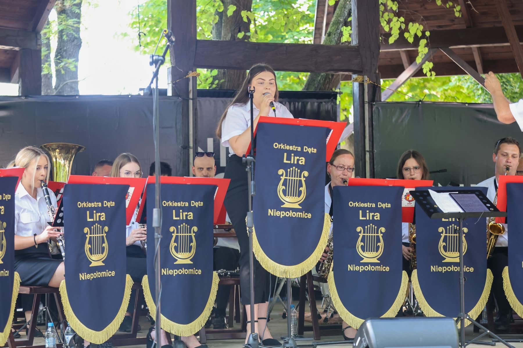 Orkiestra Dęta Lira z Niepołomic podczas występu plenerowego. Wokalistka śpiewa do mikrofonu, a muzycy grają na instrumentach dętych. Widoczne banery orkiestry.