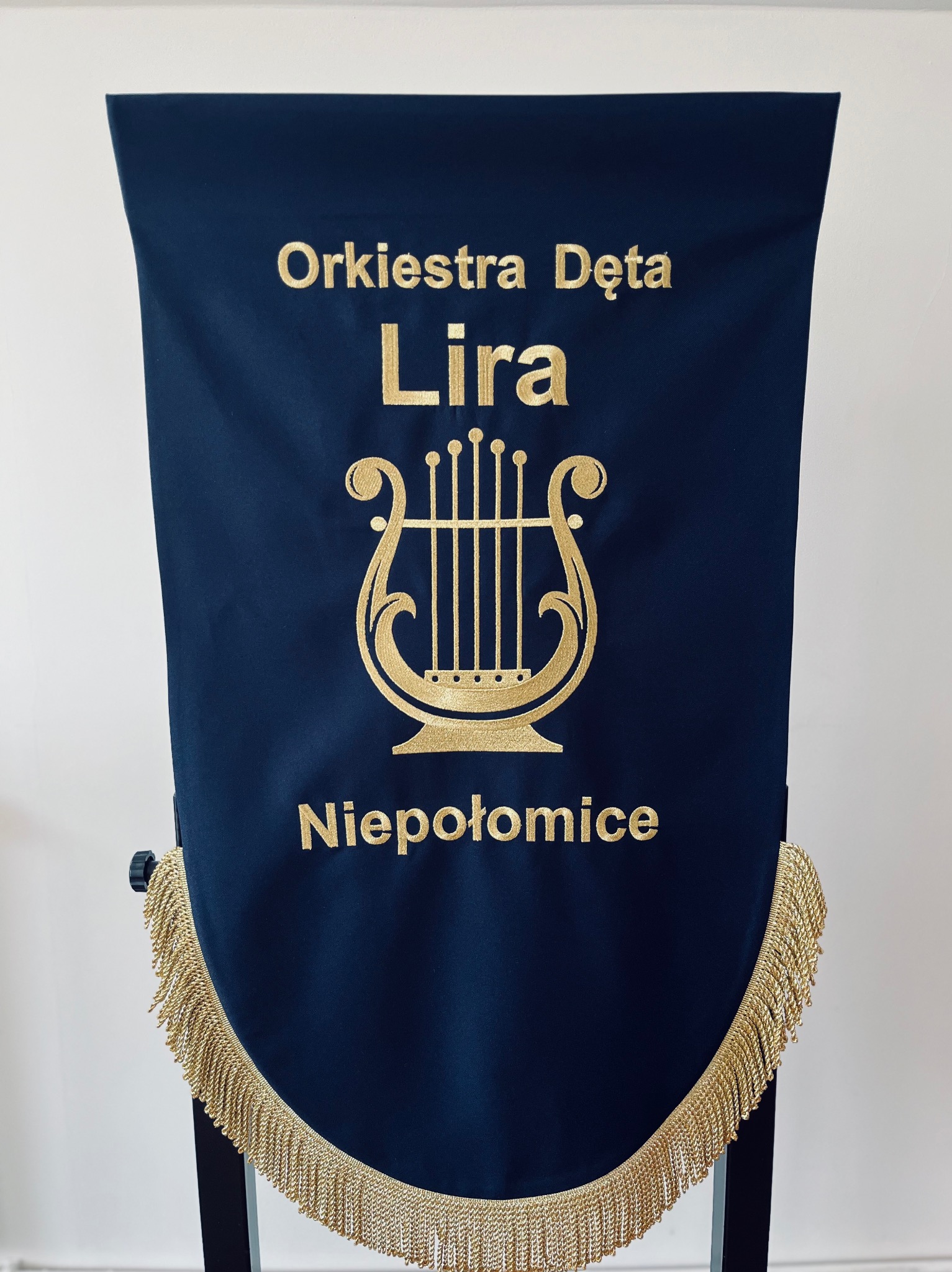 Granatowy sztandar z haftowanym złotym napisem 'Orkiestra Dęta Lira Niepołomice' i motywem liry, wykończony frędzlami. Precyzyjne wykonanie, elegancki wygląd.