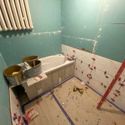 Pal-budex - Łazienka w trakcie wykończenia: częściowo ułożone białe płytki imitujące marmur na ścianie, poziomica oparta o ścianę, narzędzia na podłodze, widoczne wiadra z zaprawą i grzejnik na ścianie.