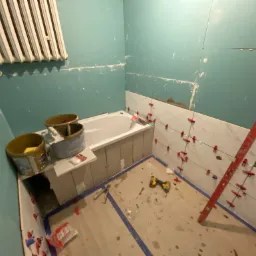 Łazienka w trakcie wykończenia: częściowo ułożone białe płytki imitujące marmur na ścianie, poziomica oparta o ścianę, narzędzia na podłodze, widoczne wiadra z zaprawą i grzejnik na ścianie.