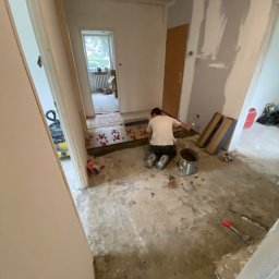 Pal-budex - Układanie płytek podłogowych w korytarzu mieszkania, widoczne narzędzia, klej, płytki i krzyżyki dystansowe, ściany częściowo przygotowane do malowania.