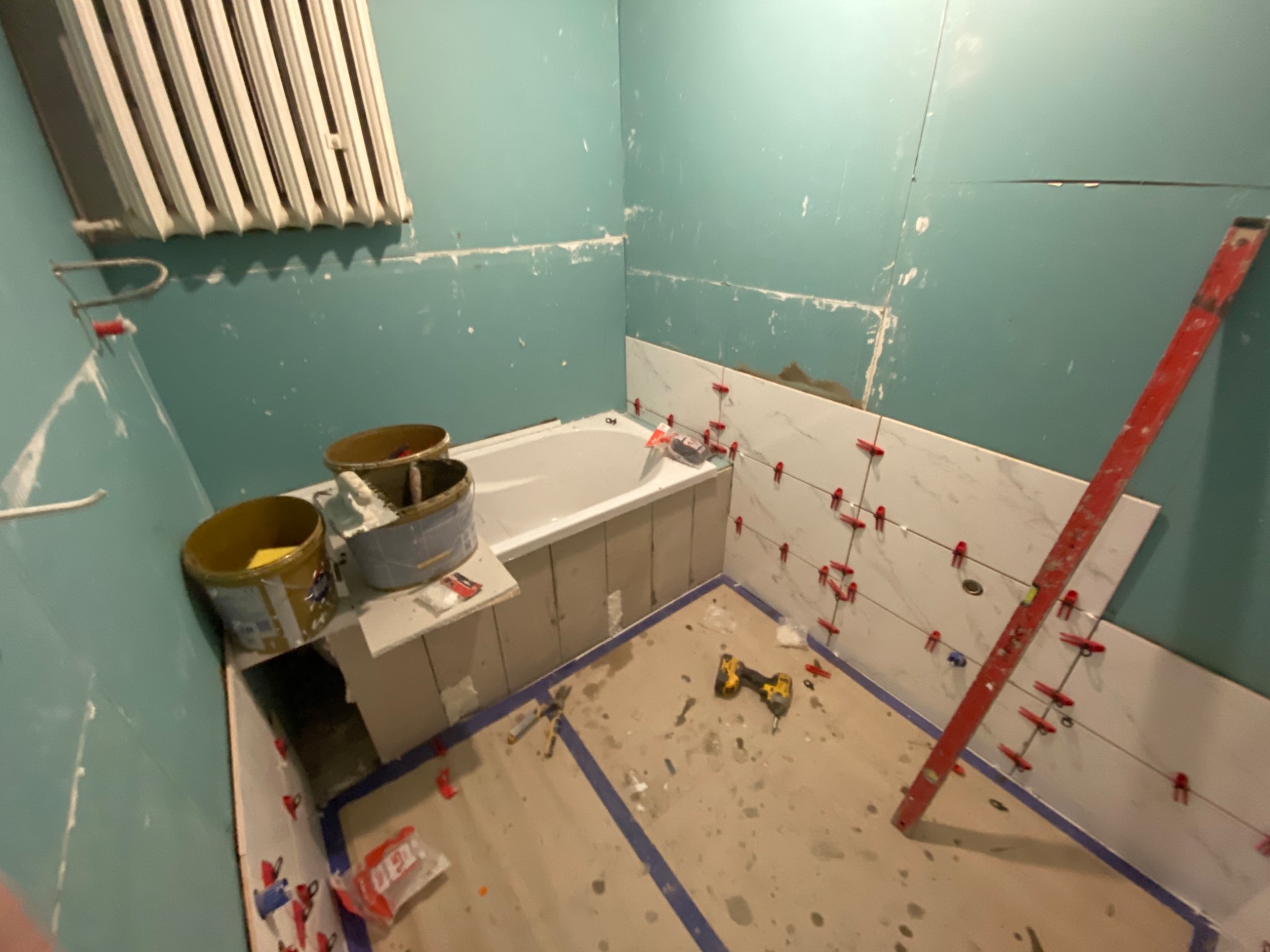 Łazienka w trakcie wykończenia: częściowo ułożone białe płytki imitujące marmur na ścianie, poziomica oparta o ścianę, narzędzia na podłodze, widoczne wiadra z zaprawą i grzejnik na ścianie.