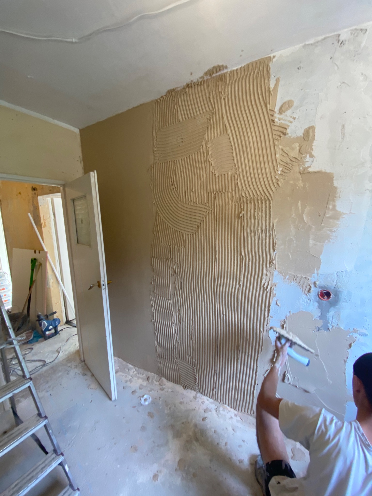 Nakładanie gładzi szpachlowej na ścianę w trakcie remontu mieszkania, widoczne ślady po gładzi na podłodze, drabina i narzędzia w tle.