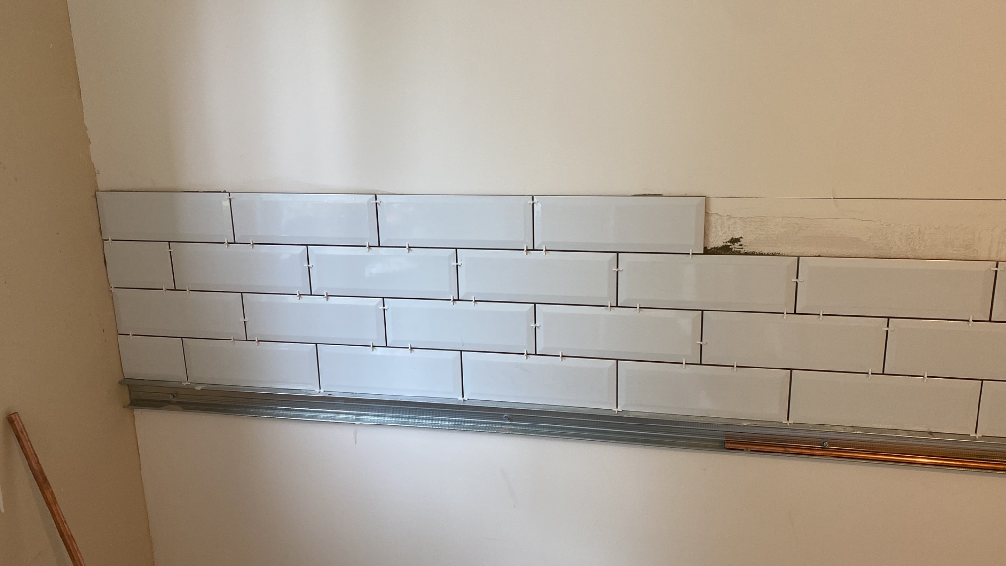 Układanie błyszczących, białych płytek typu metro na ścianie; widoczna listwa montażowa i krzyżyki dystansowe, fragment ściany bez glazury.