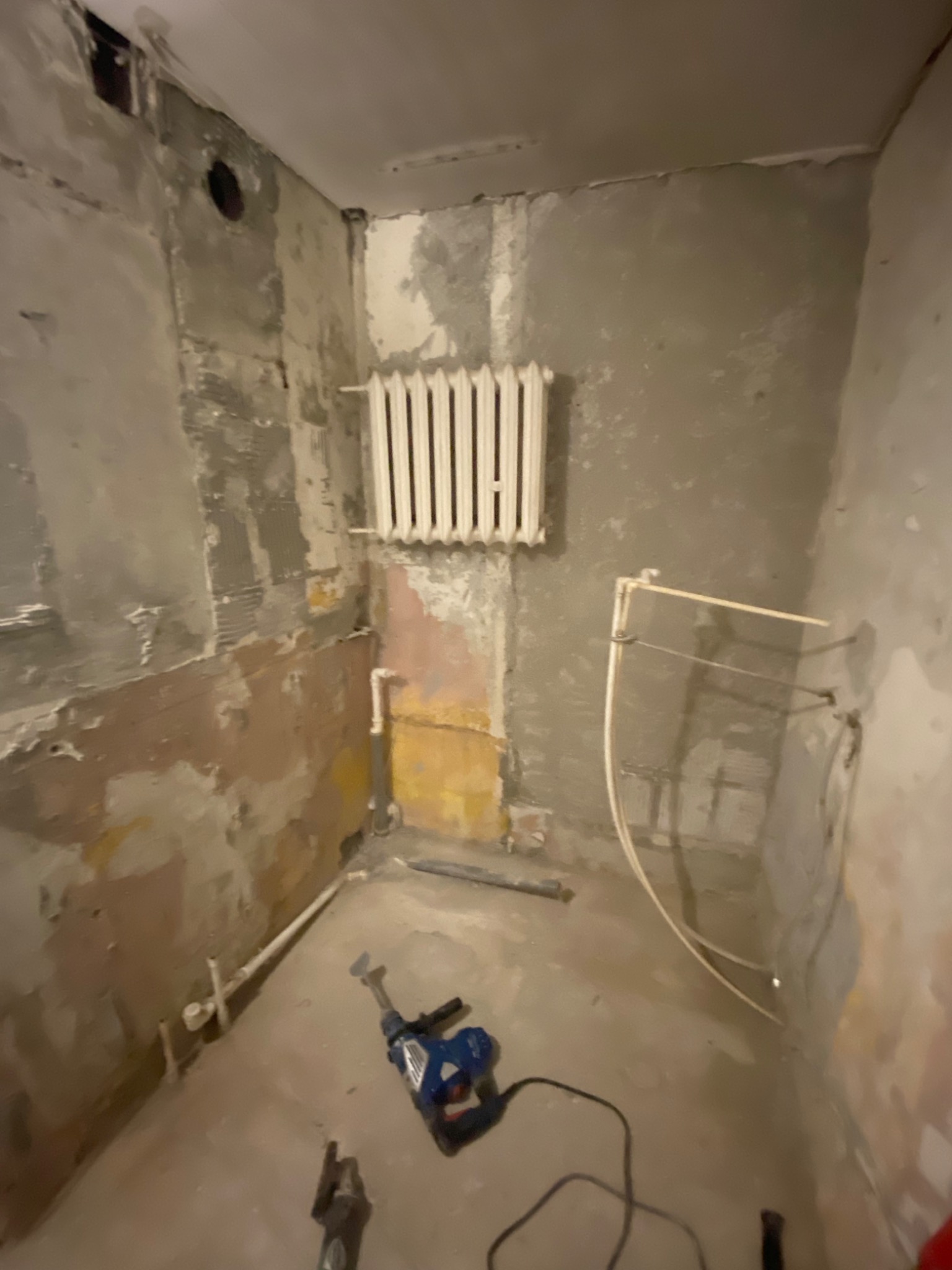 Wnętrze łazienki w trakcie demontażu starych płytek, widoczny grzejnik, rury, ściany w stanie surowym i młot udarowy leżący na podłodze.