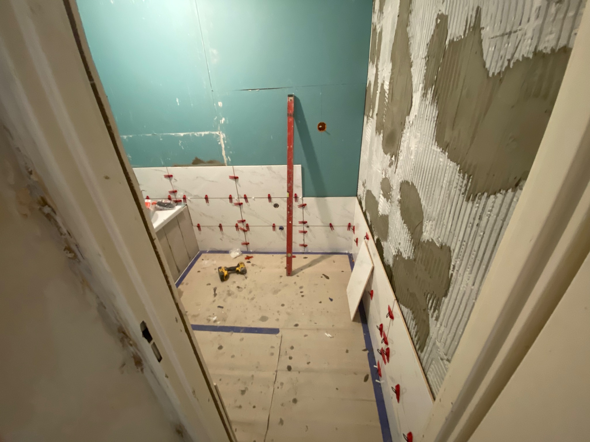 Łazienka w trakcie układania białych płytek marmurowych na ścianach, widoczne poziomica, wiertarka akumulatorowa, krzyżyki dystansowe i ślady zaprawy klejowej na ścianie.