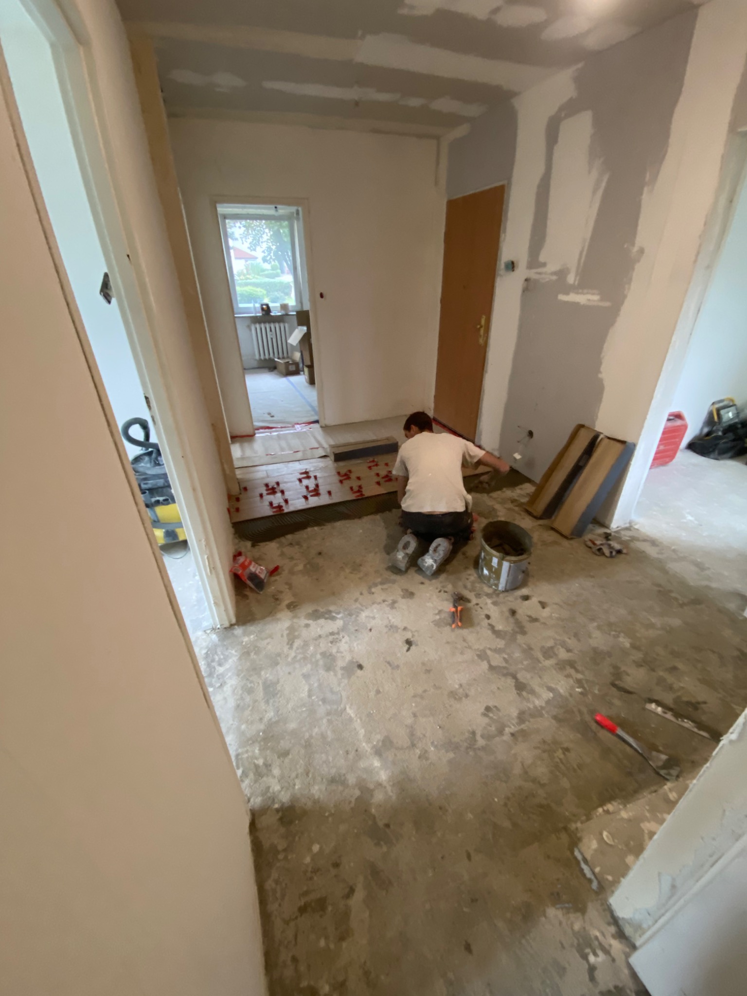 Układanie płytek podłogowych w korytarzu mieszkania, widoczne narzędzia, klej, płytki i krzyżyki dystansowe, ściany częściowo przygotowane do malowania.