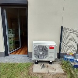 Chłodnictwo i klimatyzacja - Zewnętrzna jednostka klimatyzacyjna marki Vivax umieszczona na betonowej podstawie obok ściany budynku i otwartego okna tarasowego z widocznym wnętrzem, przylegająca do trawnika i ogrodzenia...