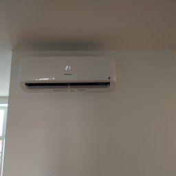 Chłodnictwo i klimatyzacja - Biały klimatyzator Hisense zamontowany na białej ścianie, wyświetlający temperaturę 21 stopni Celsjusza, widok z dołu w jasnym pomieszczeniu z oknem.