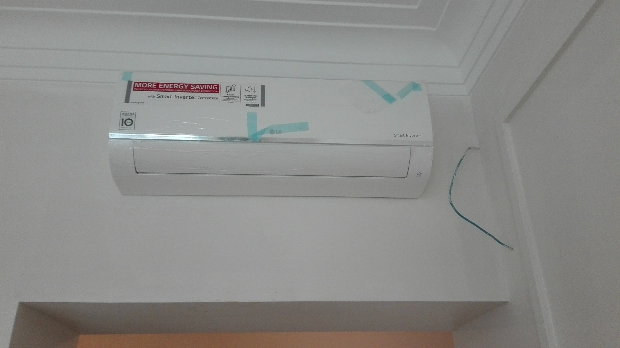 Nowy, biały klimatyzator LG Smart Inverter, jeszcze z folią ochronną i taśmą zabezpieczającą, zamontowany na białej ścianie w pomieszczeniu. Widoczne wystające przewody elektryczne.