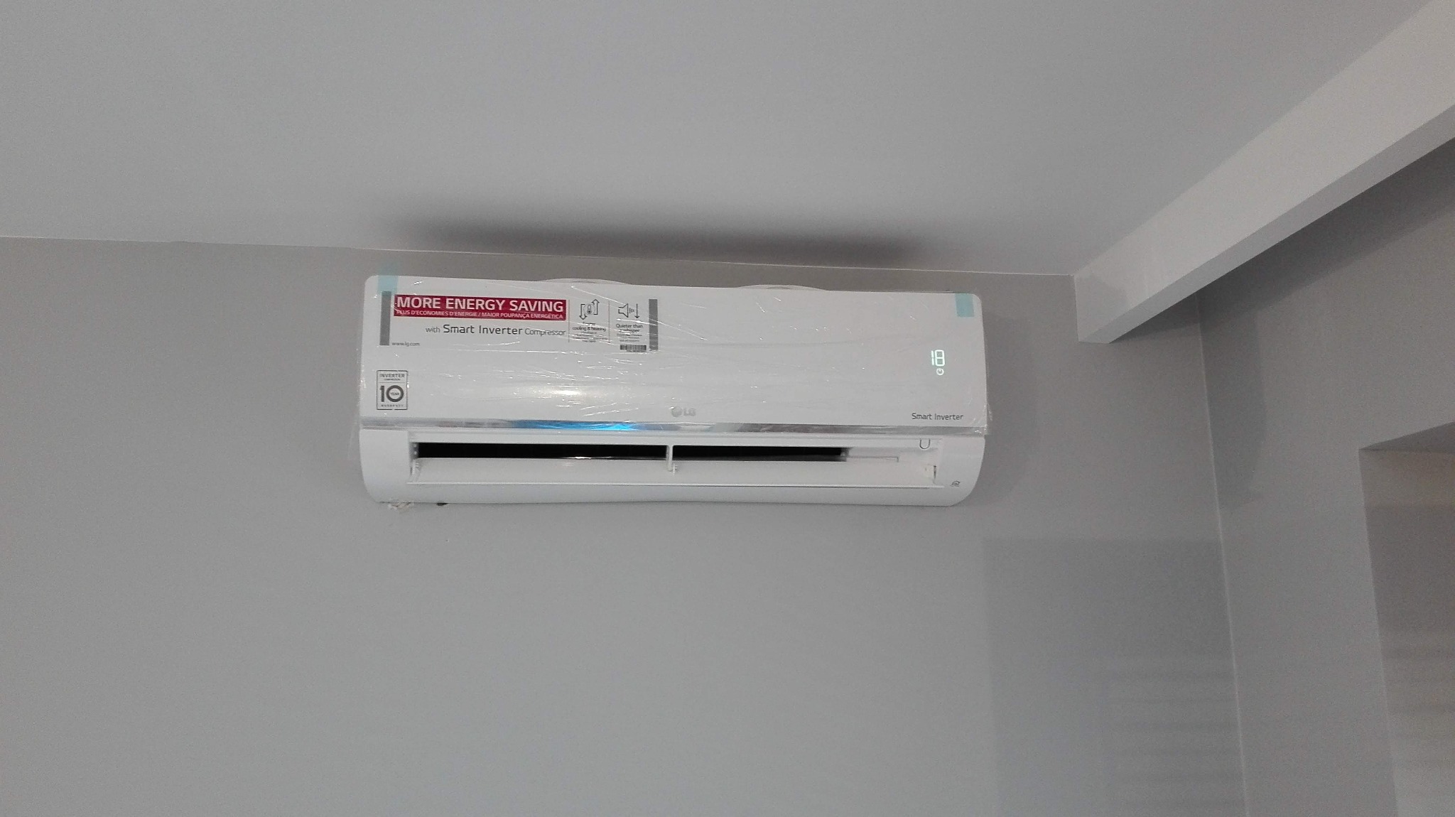 Nowy, biały klimatyzator LG Smart Inverter zamontowany na szarej ścianie, jeszcze w folii ochronnej, wyświetlający temperaturę 18 stopni Celsjusza.
