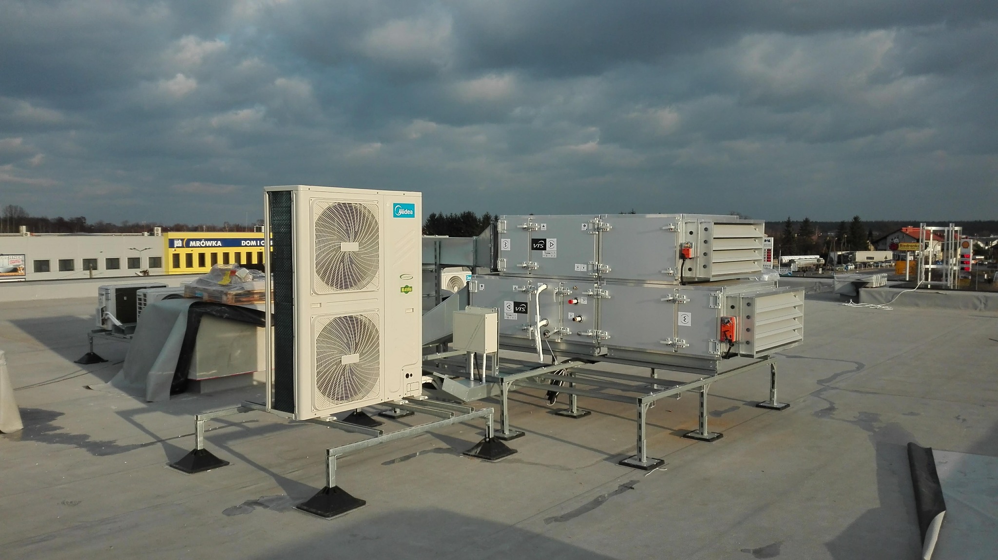 Klimatyzacja przemysłowa Midea z dwoma wentylatorami oraz modułowa centrala wentylacyjna VIS zamontowane na dachu budynku z widokiem na sklep Mrówka w tle.